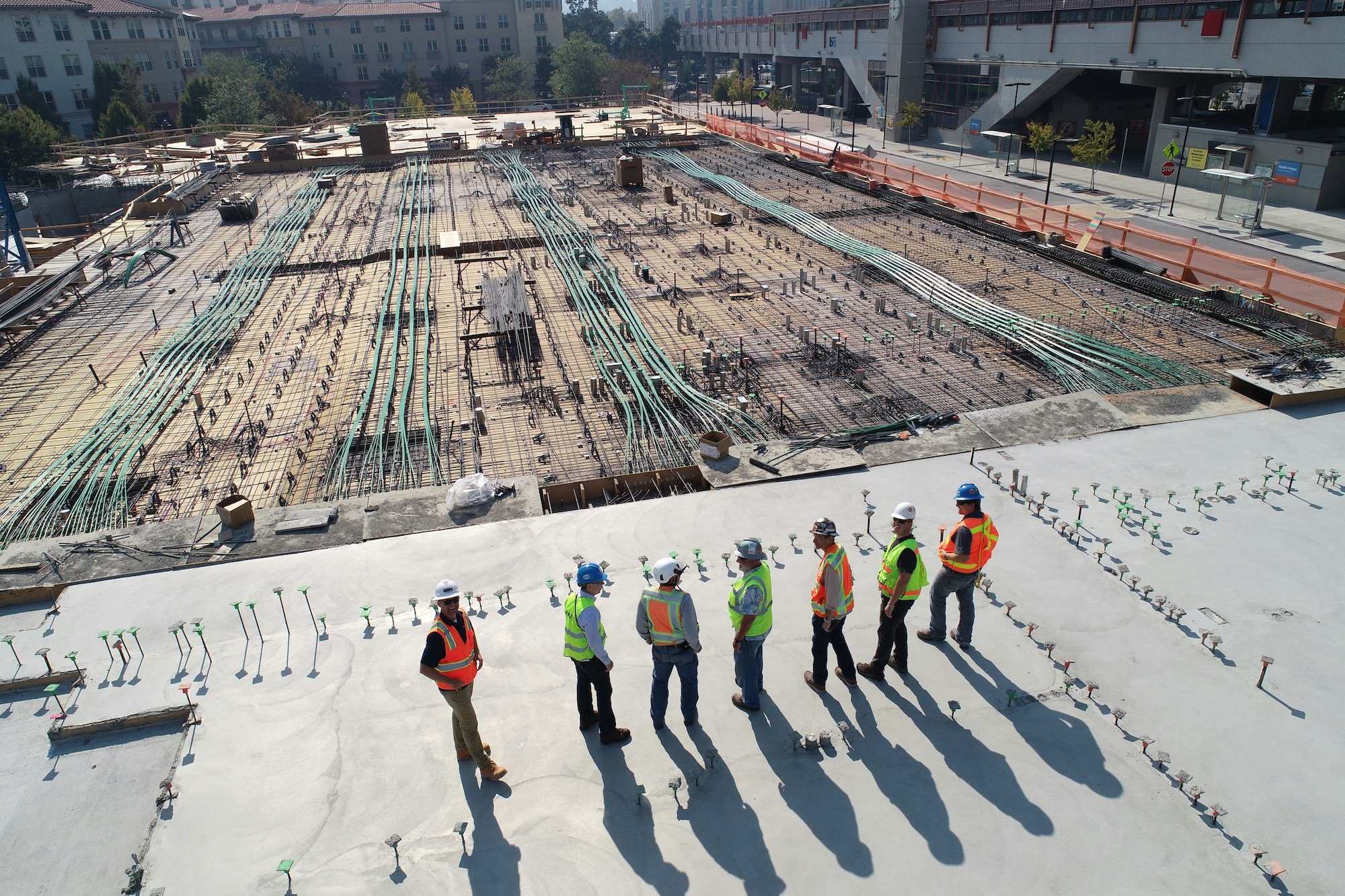 Seven construction workers in safety vests and helmets standing on a concrete slab at a building site.