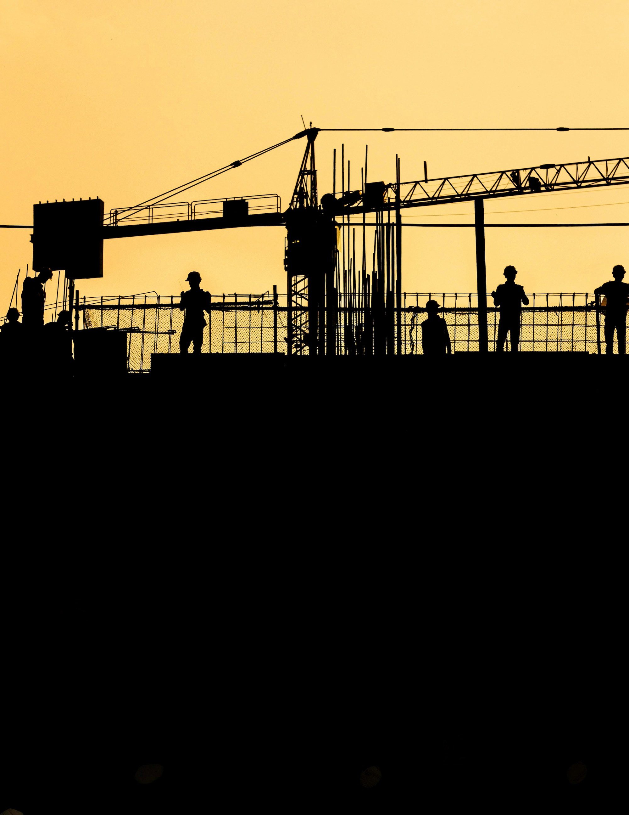 Silhouettes of construction workers and a crane against an orange sky at a building site.