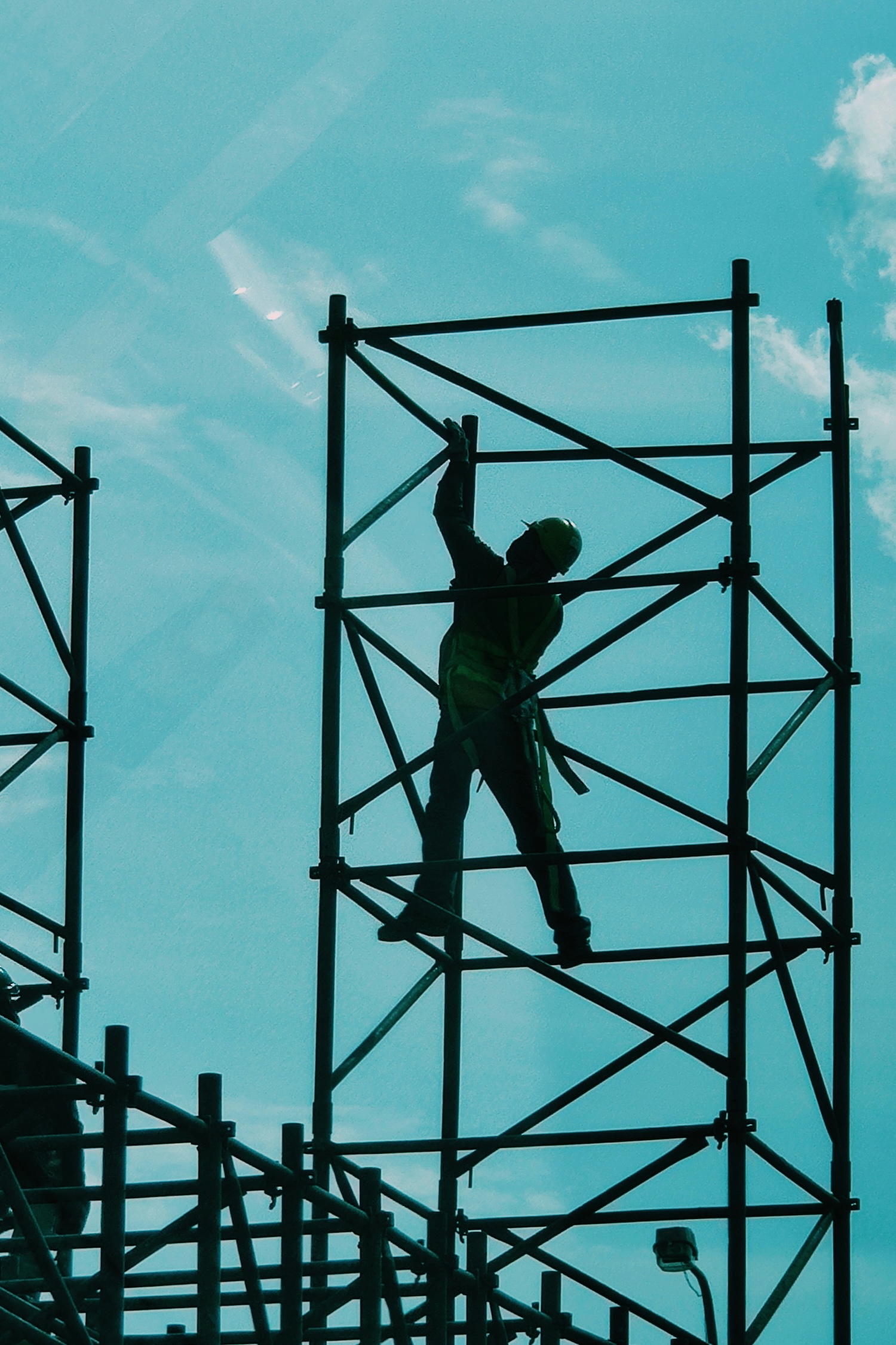 Silhouette of a construction worker wearing a safety harness and helmet on metal scaffolding against blue sky.