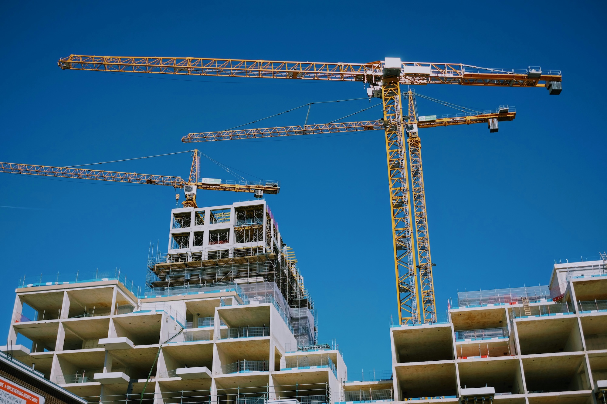 Concrete building under construction with three large yellow tower cranes against a clear blue sky.