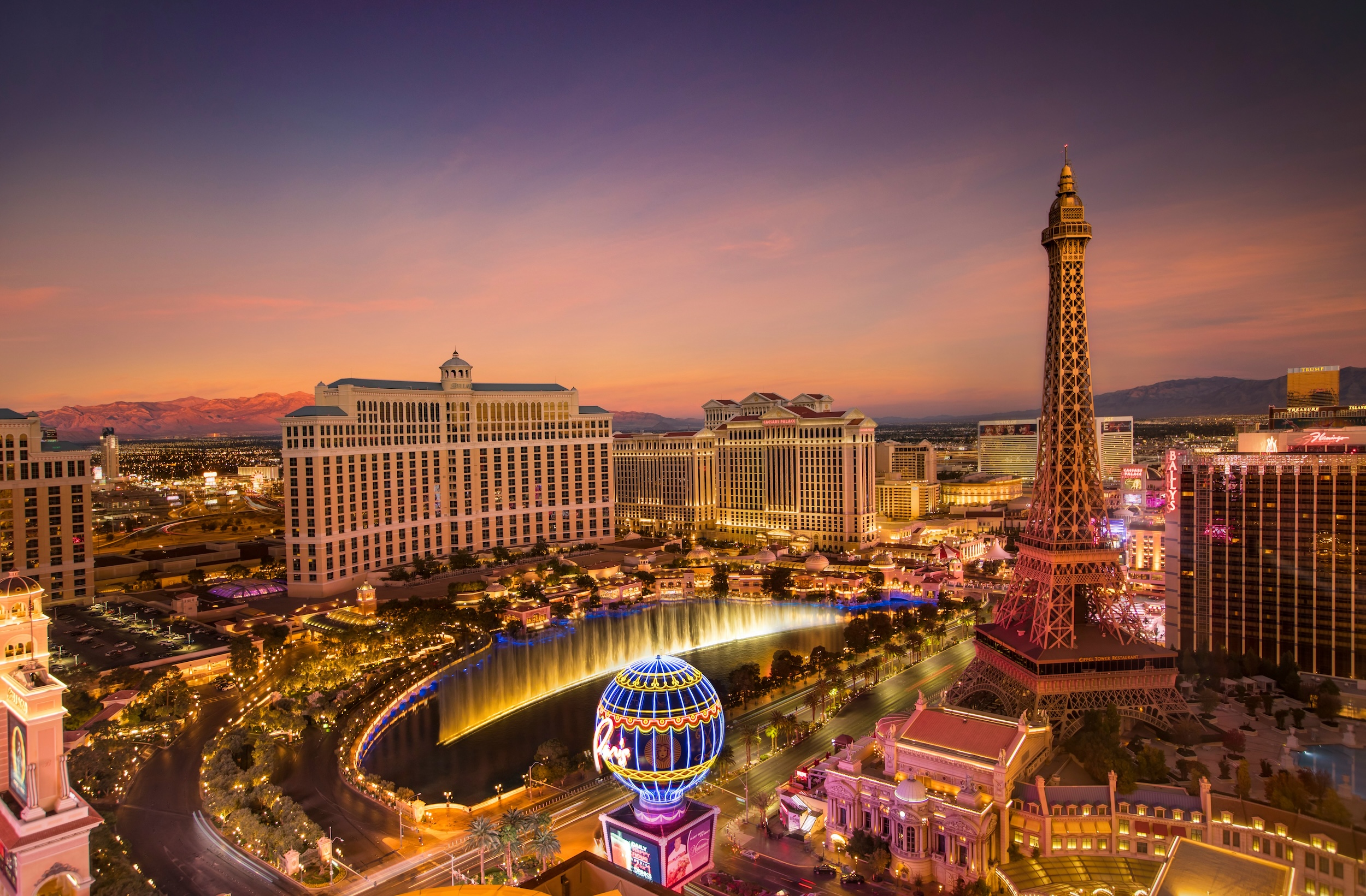 Las Vegas Strip at sunset with Bellagio fountains, Paris Eiffel Tower, and surrounding hotels lit up.