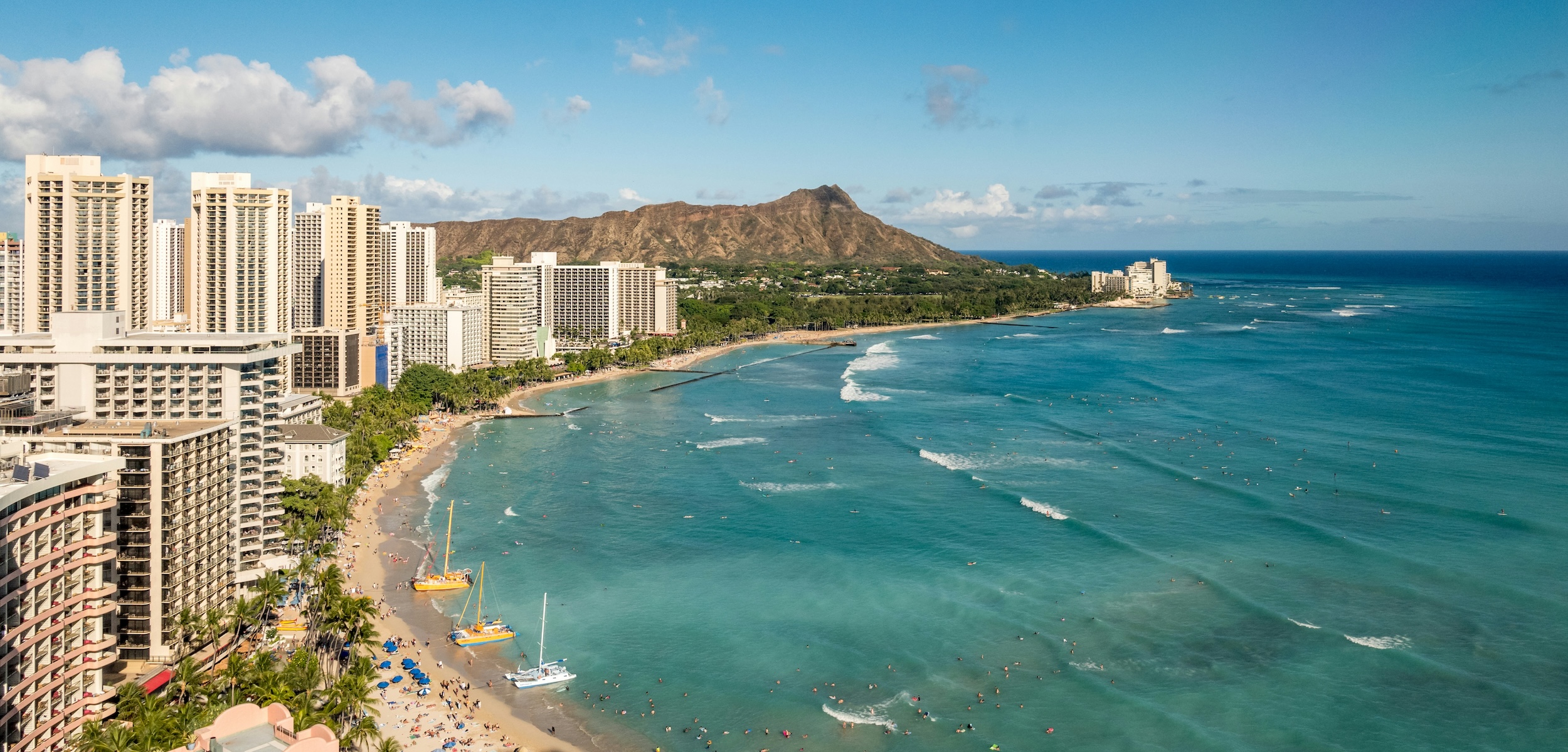 Aerial view of Waikiki Beach with high-rise hotels, blue ocean, surfers, and Diamond Head crater in the background.