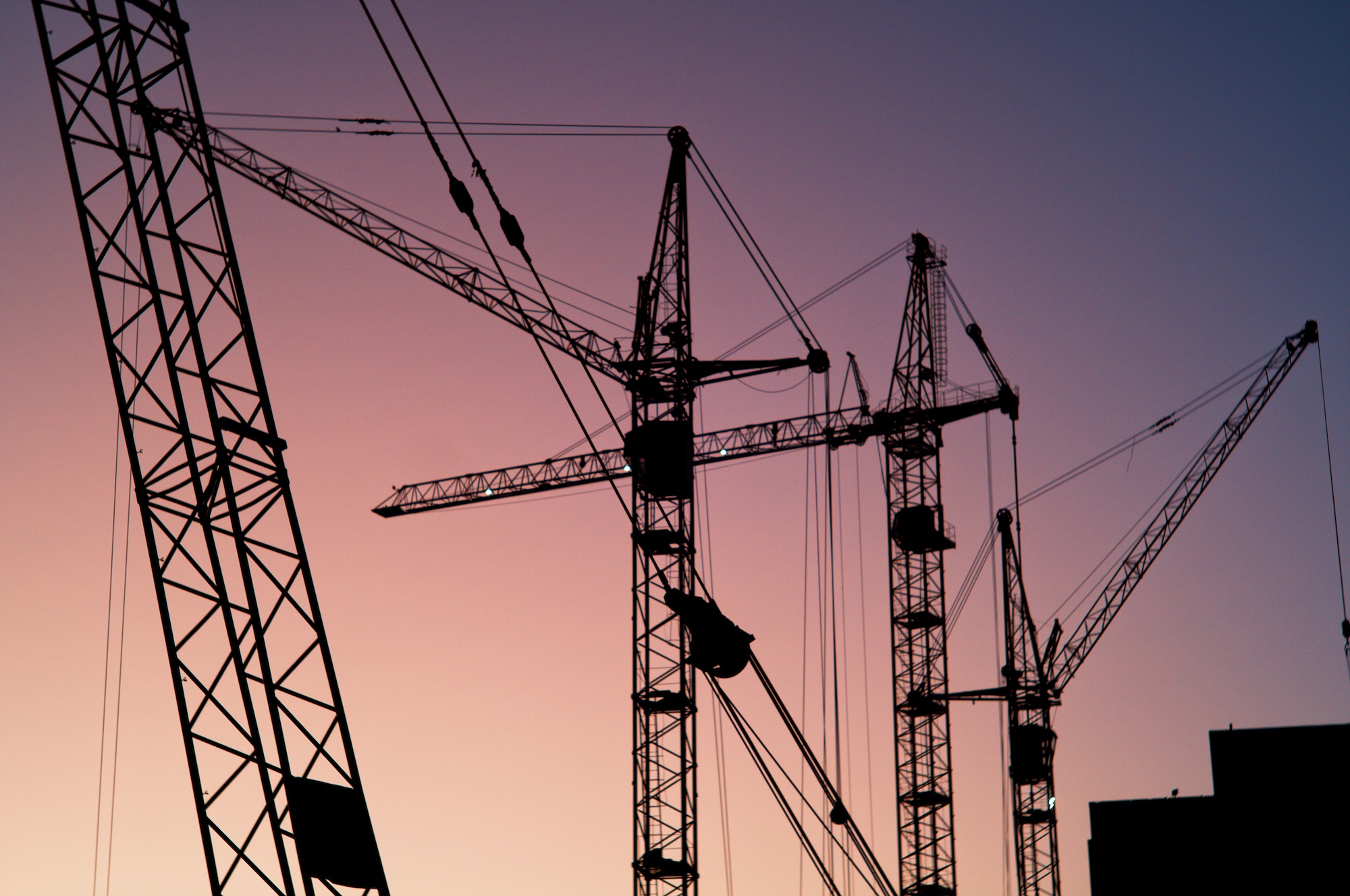 Silhouettes of construction cranes against a pink and purple sunset sky.