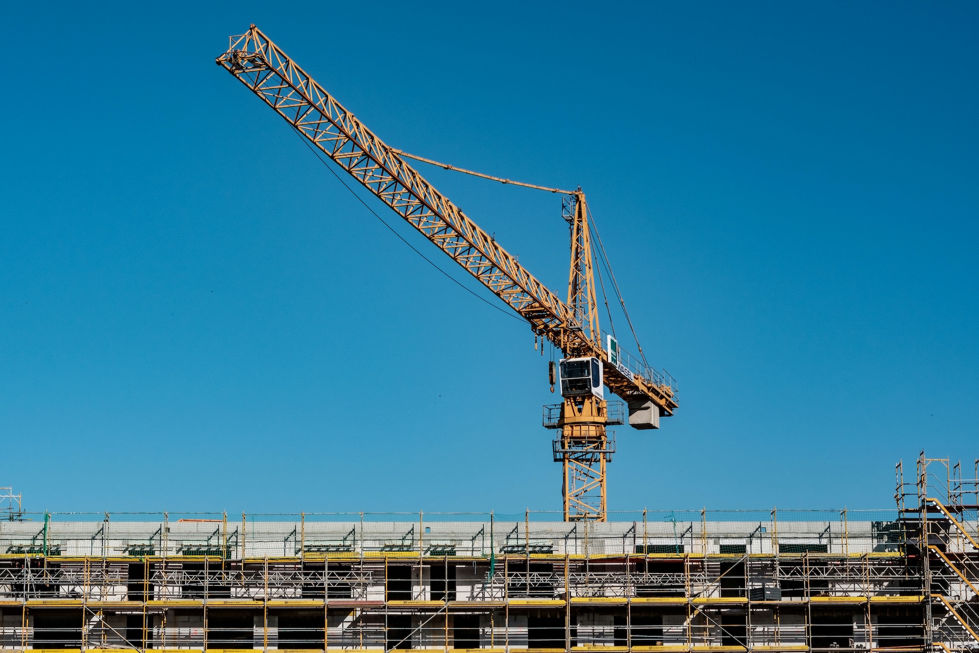 Yellow construction crane towering over a building under construction with scaffolding on a clear blue sky.