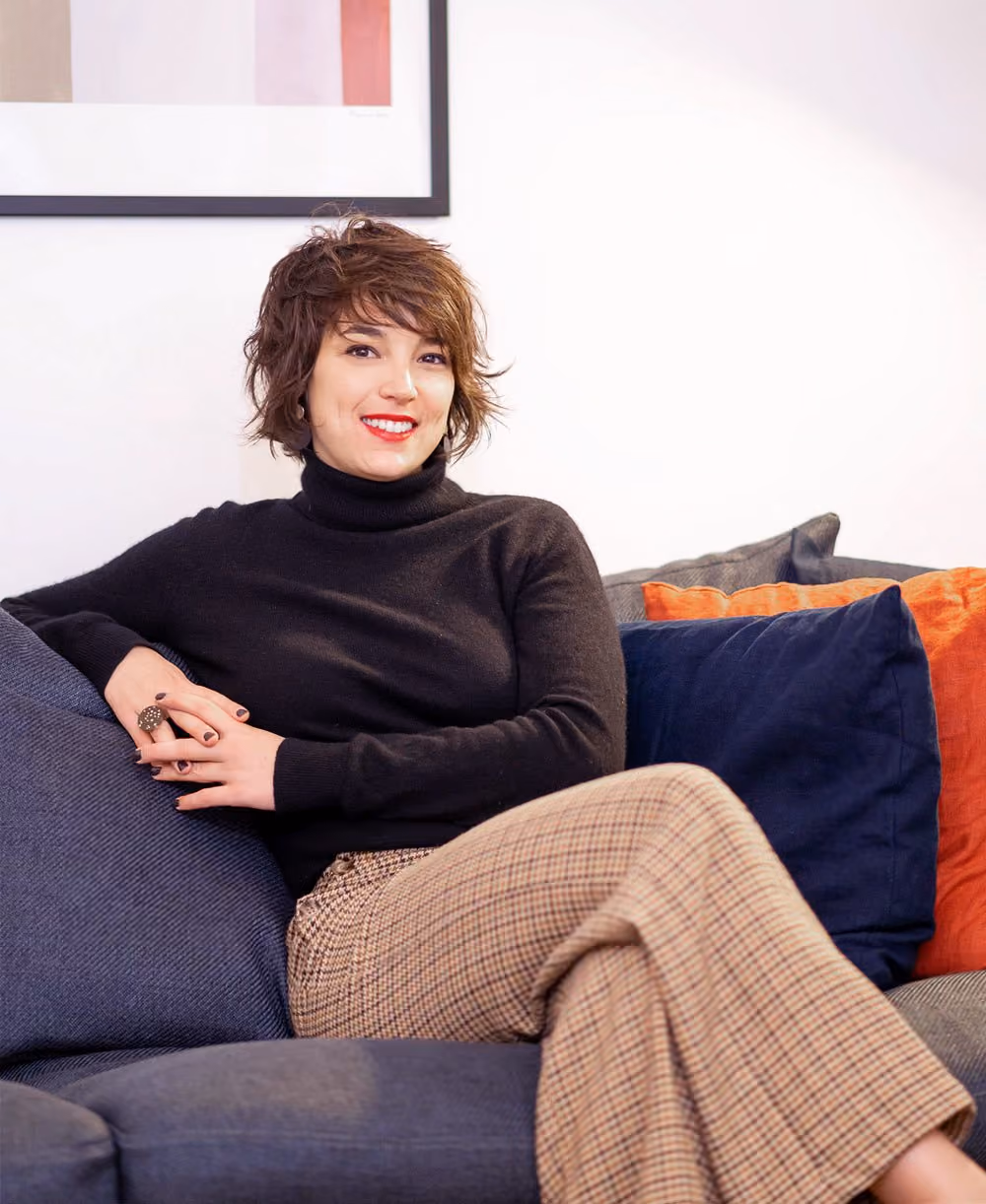 Femme souriante en pull noir et pantalon à carreaux assise sur un canapé avec des coussins colorés.