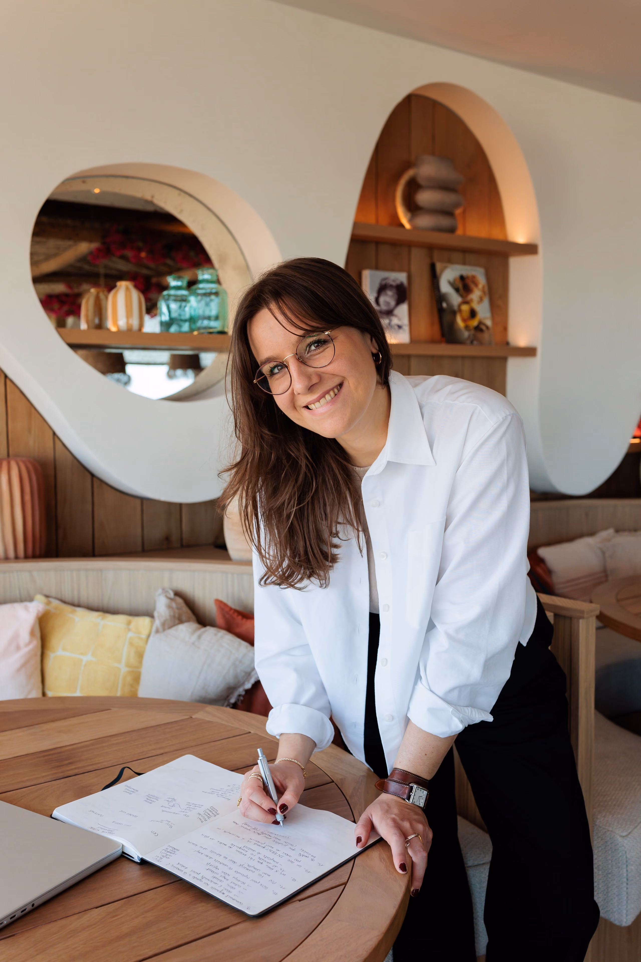 Femme souriante écrivant dans un carnet posé sur une table en bois dans un salon cosy.