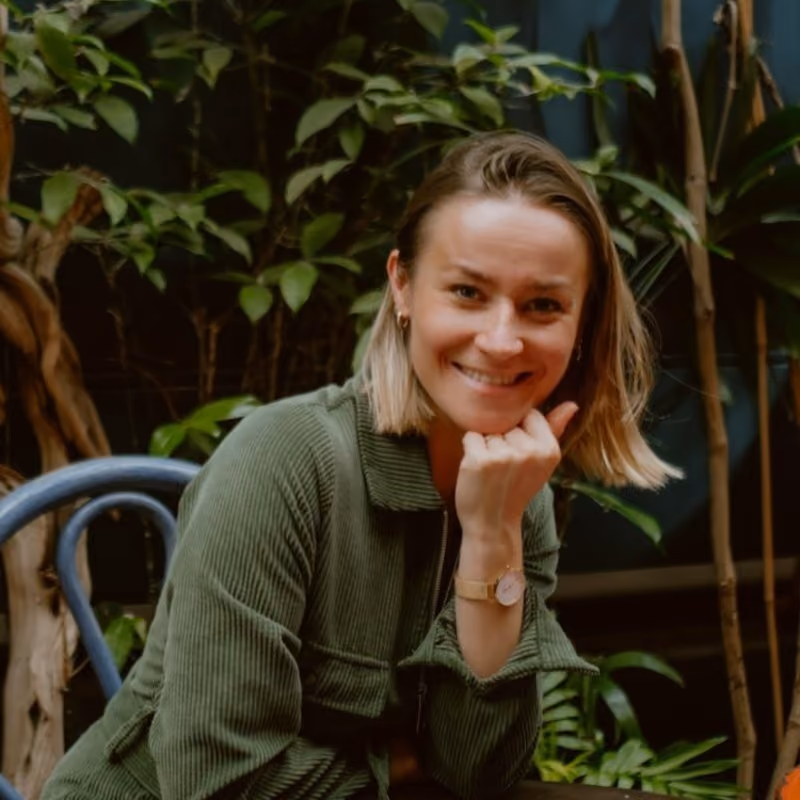 Femme souriante en veste verte assise sur une chaise dans un décor végétal intérieur.