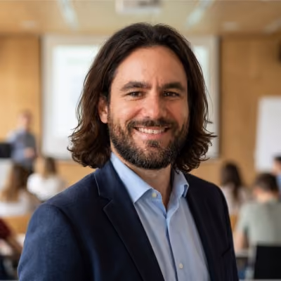 Homme souriant aux cheveux mi-longs et barbe portant un costume dans une salle de classe floue.