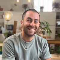 Un homme souriant aux cheveux courts porte un t-shirt gris, assis dans un intérieur avec plantes et meubles.