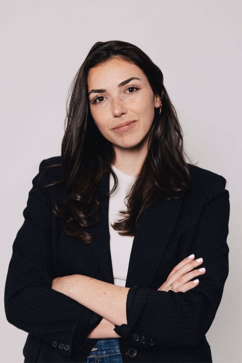 Portrait d'une femme aux cheveux longs bruns, bras croisés, en veste noire sur fond clair.
