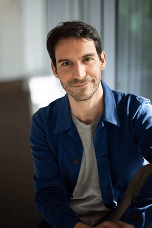 Homme souriant portant une veste bleue et un t-shirt gris, assis devant un fond flou.