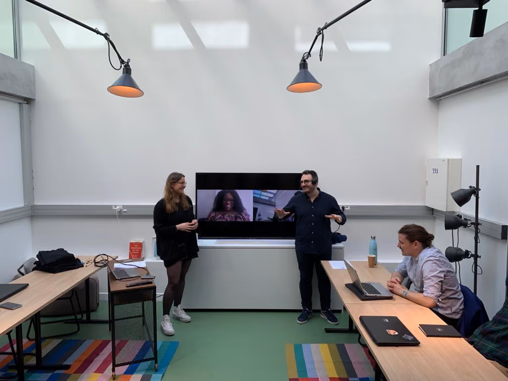 Trois personnes en réunion dans une salle lumineuse avec deux participants en visioconférence à l'écran.
