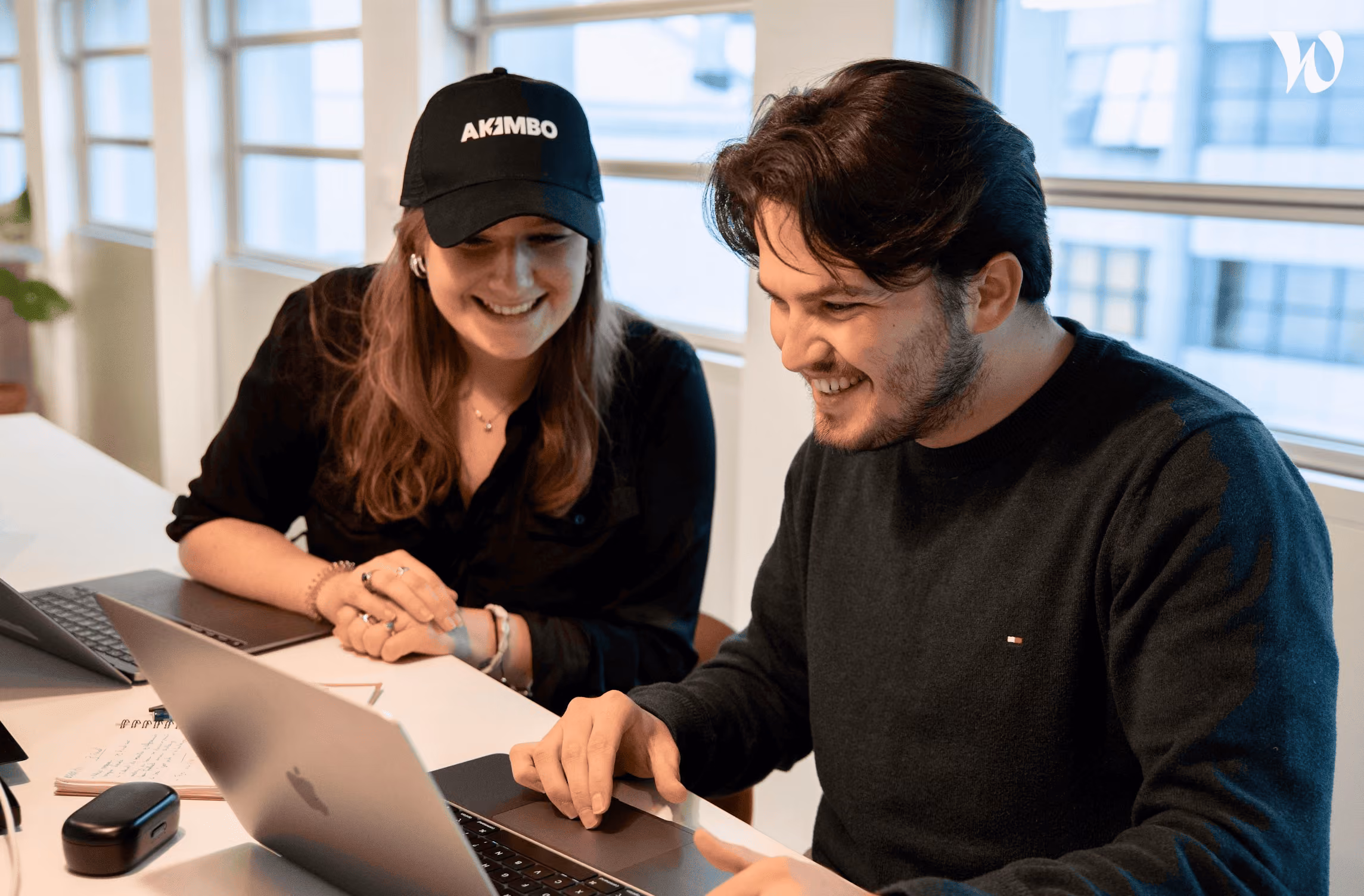 Deux collègues souriants travaillent ensemble sur un ordinateur portable dans un bureau lumineux.