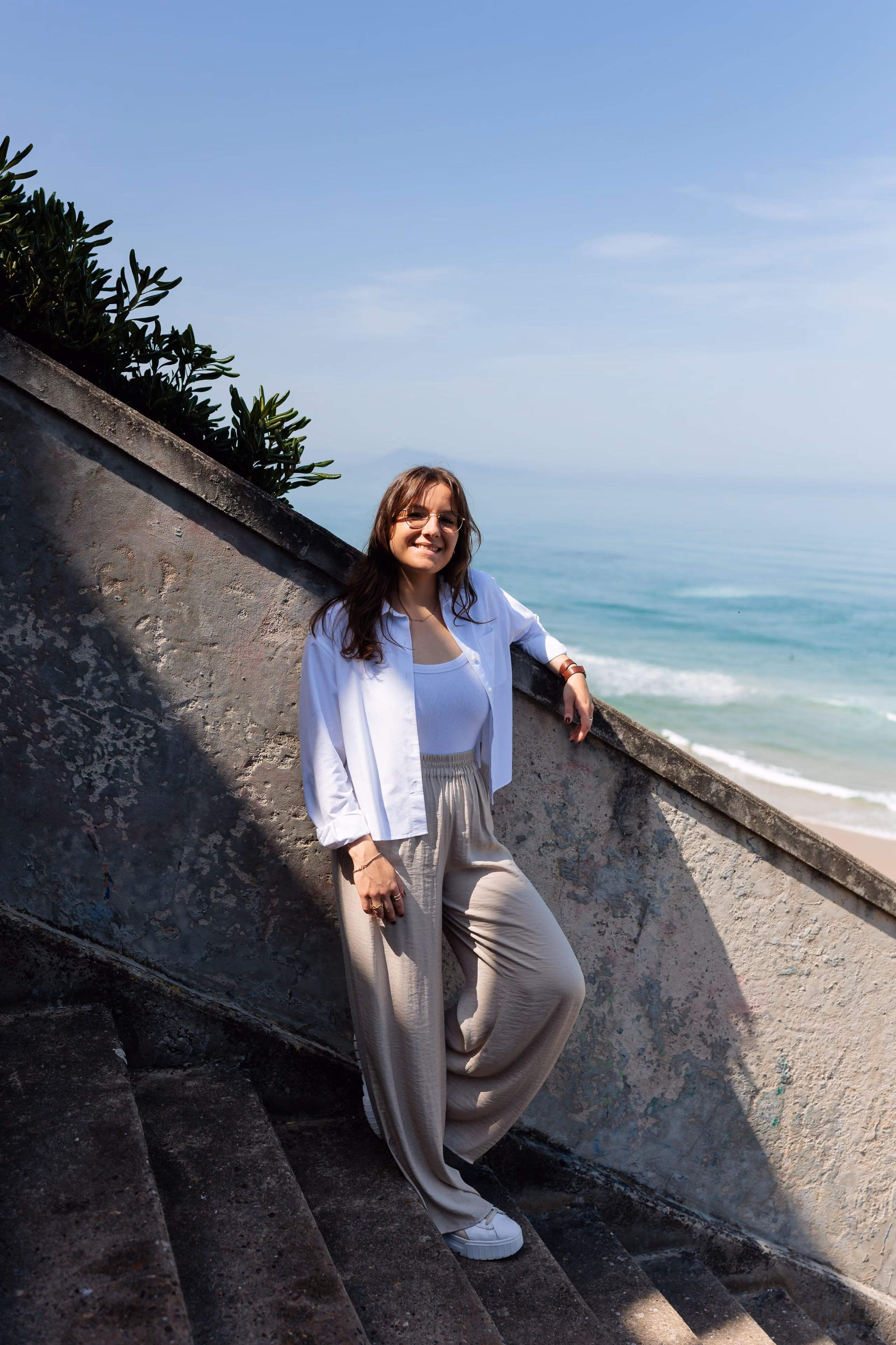 Jeune femme souriante appuyée sur un mur d'escalier avec vue sur la mer en arrière-plan.