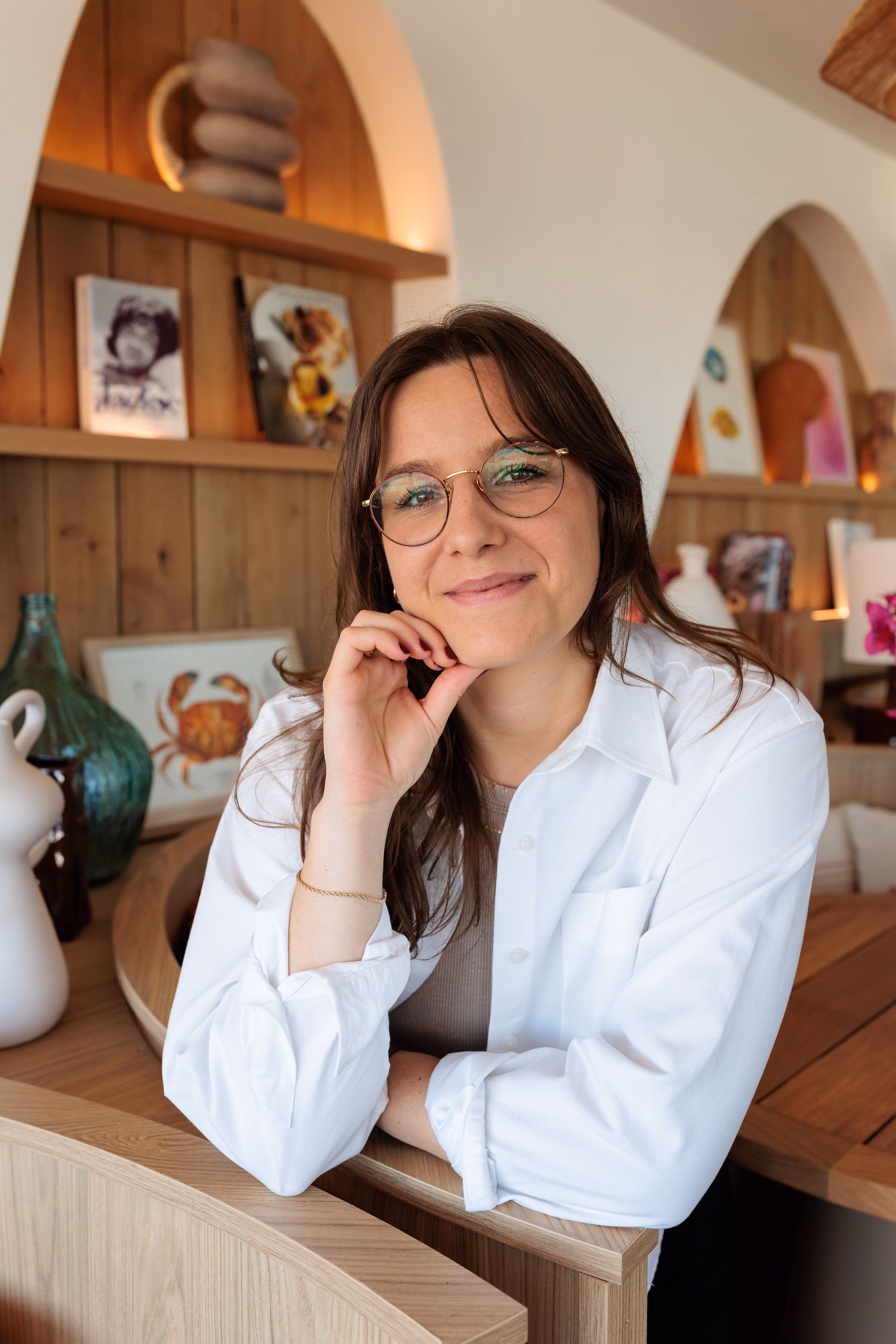 Jeune femme portant des lunettes souriant, appuyée sur un comptoir en bois dans un intérieur cosy.