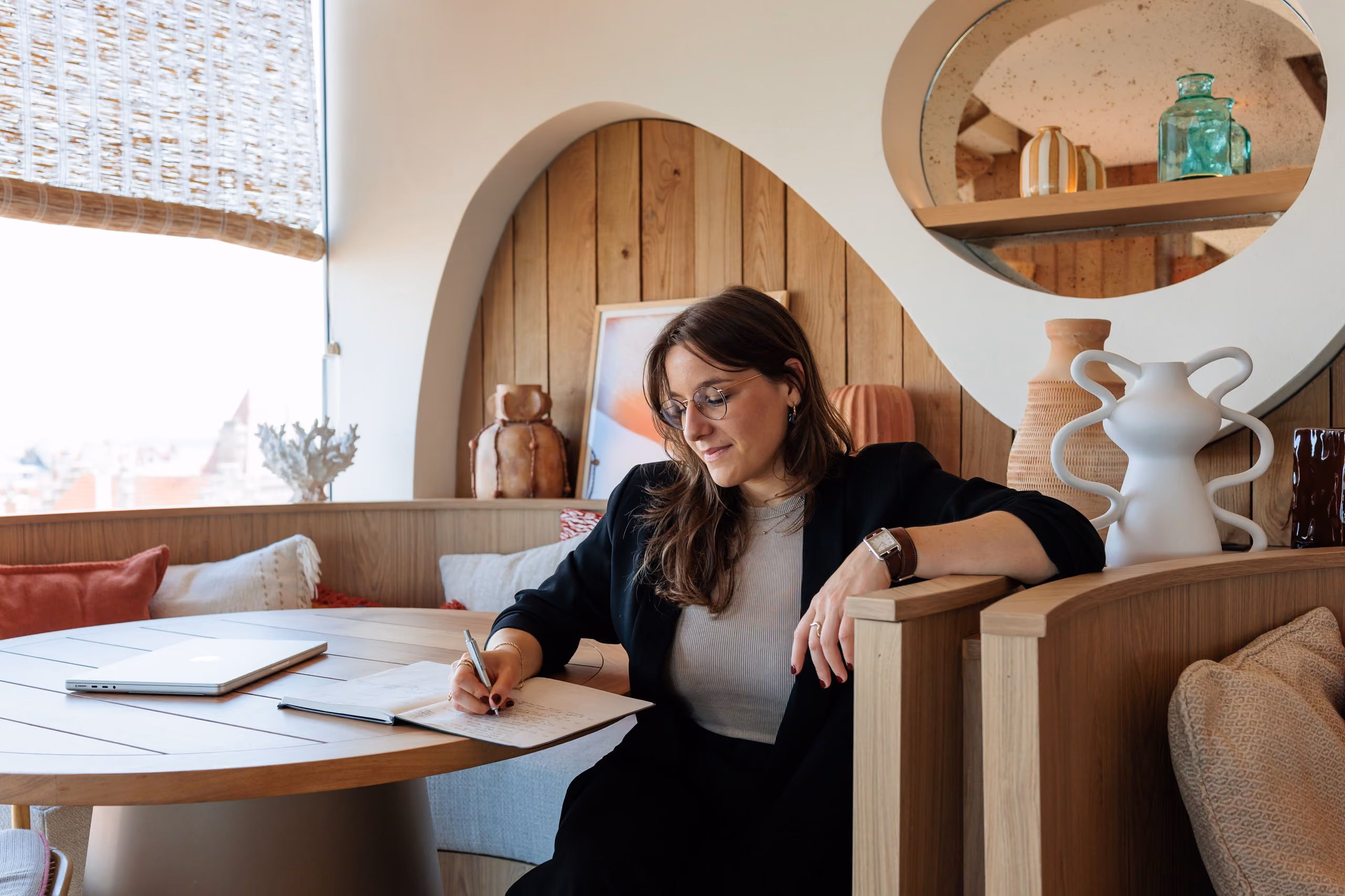 Femme écrivant dans un carnet assise à une table en bois dans un salon décoré moderne et confortable.