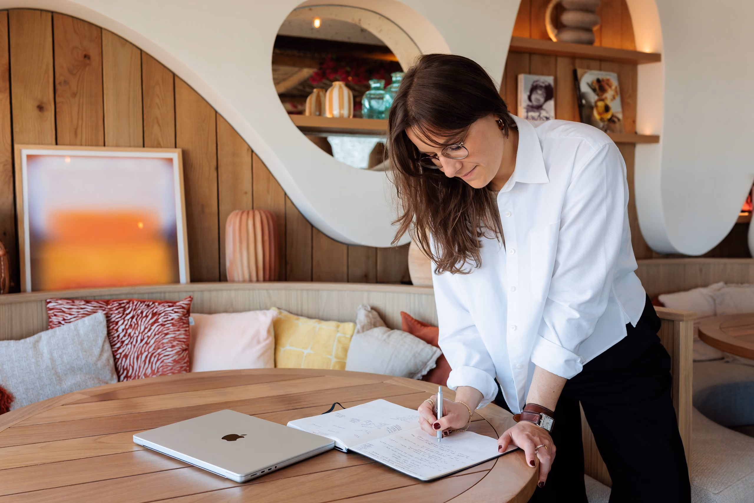 Une femme en chemise blanche écrit dans un carnet posé sur une table ronde en bois avec un ordinateur portable fermé.