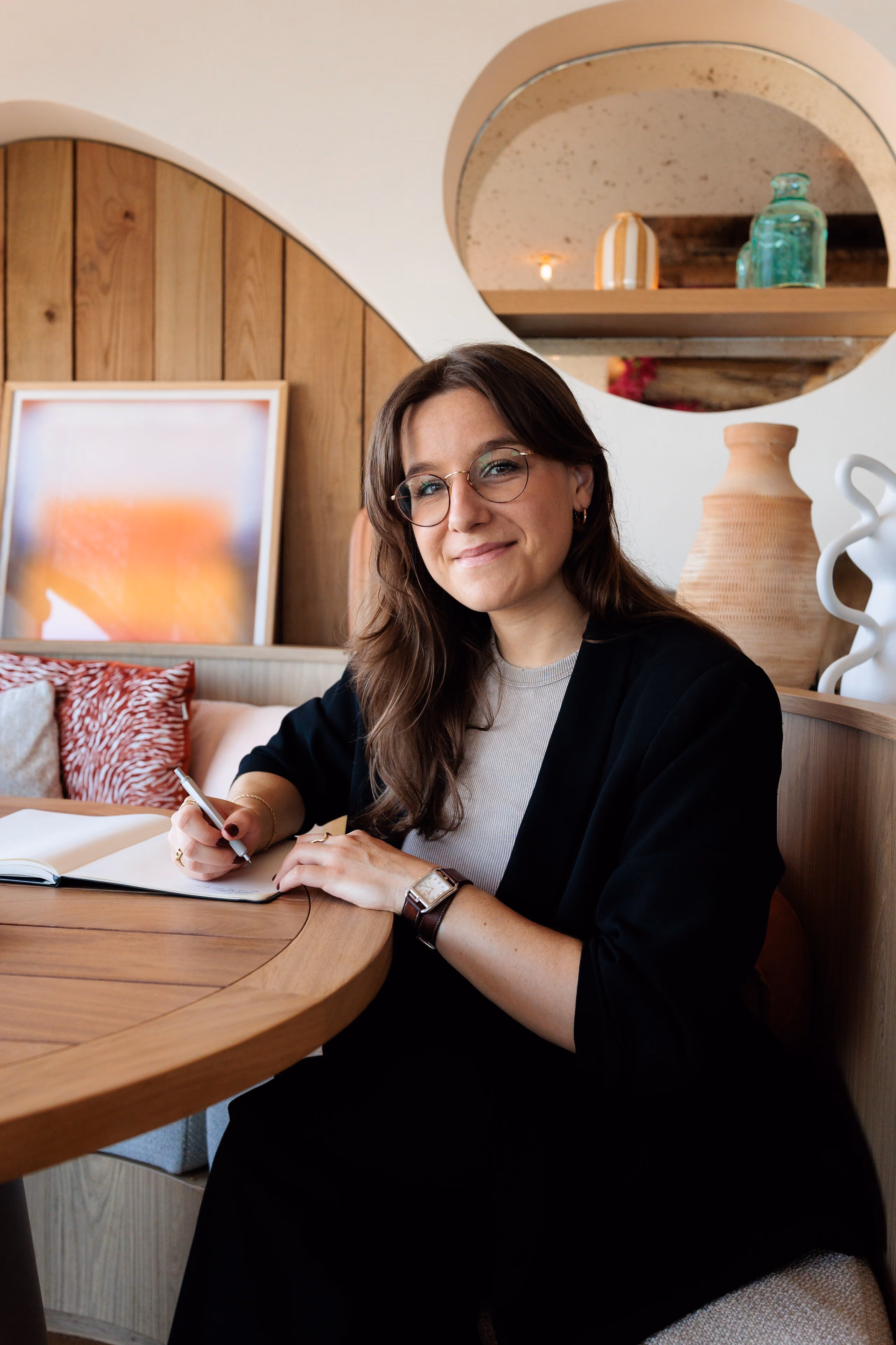 Femme souriante avec lunettes écrivant dans un carnet assise à une table en bois dans un décor moderne.