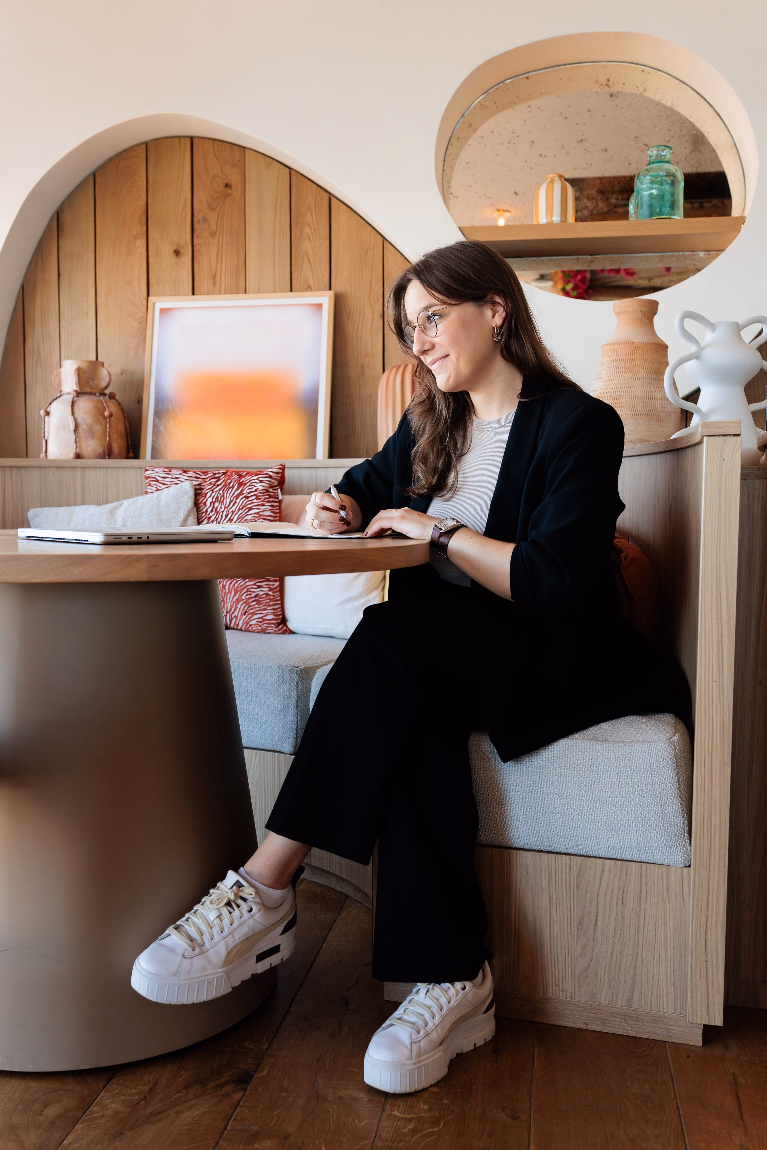 Une femme souriante portant des lunettes écrit dans un carnet assise dans un coin salon moderne.