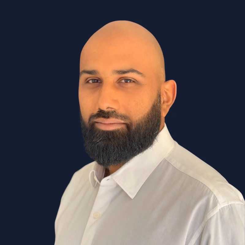 Homme chauve avec barbe dense portant une chemise blanche sur fond bleu foncé.