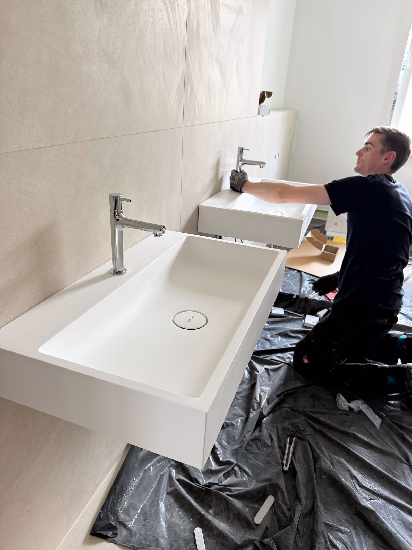 Ein Clara Mitarbeiter montiert zwei weiße Wandwaschbecken mit verchromten Wasserhähnen in einem modernen Badezimmer.