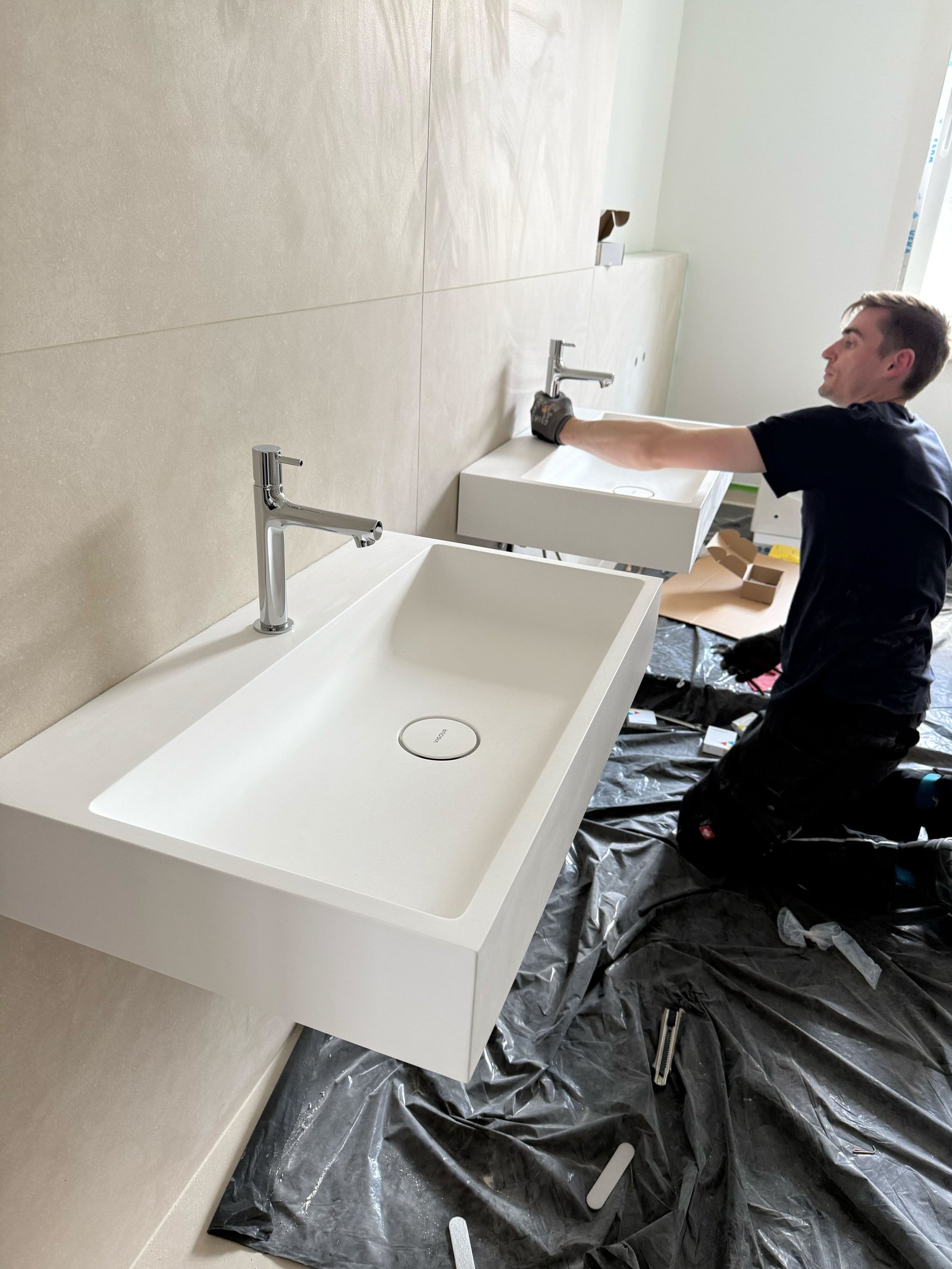 Ein Clara Mitarbeiter montiert zwei weiße Wandwaschbecken mit verchromten Wasserhähnen in einem modernen Badezimmer.