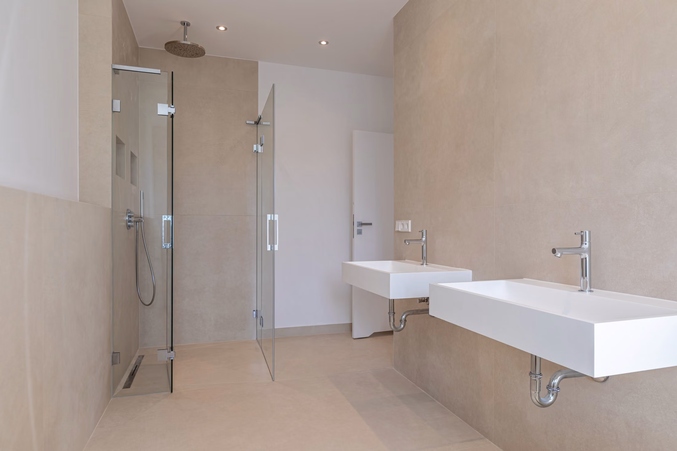 Minimalistisches, beiges Badezimmer mit begehbarer Glasdusche und zwei weißen Wandwaschbecken mit Chromarmaturen.