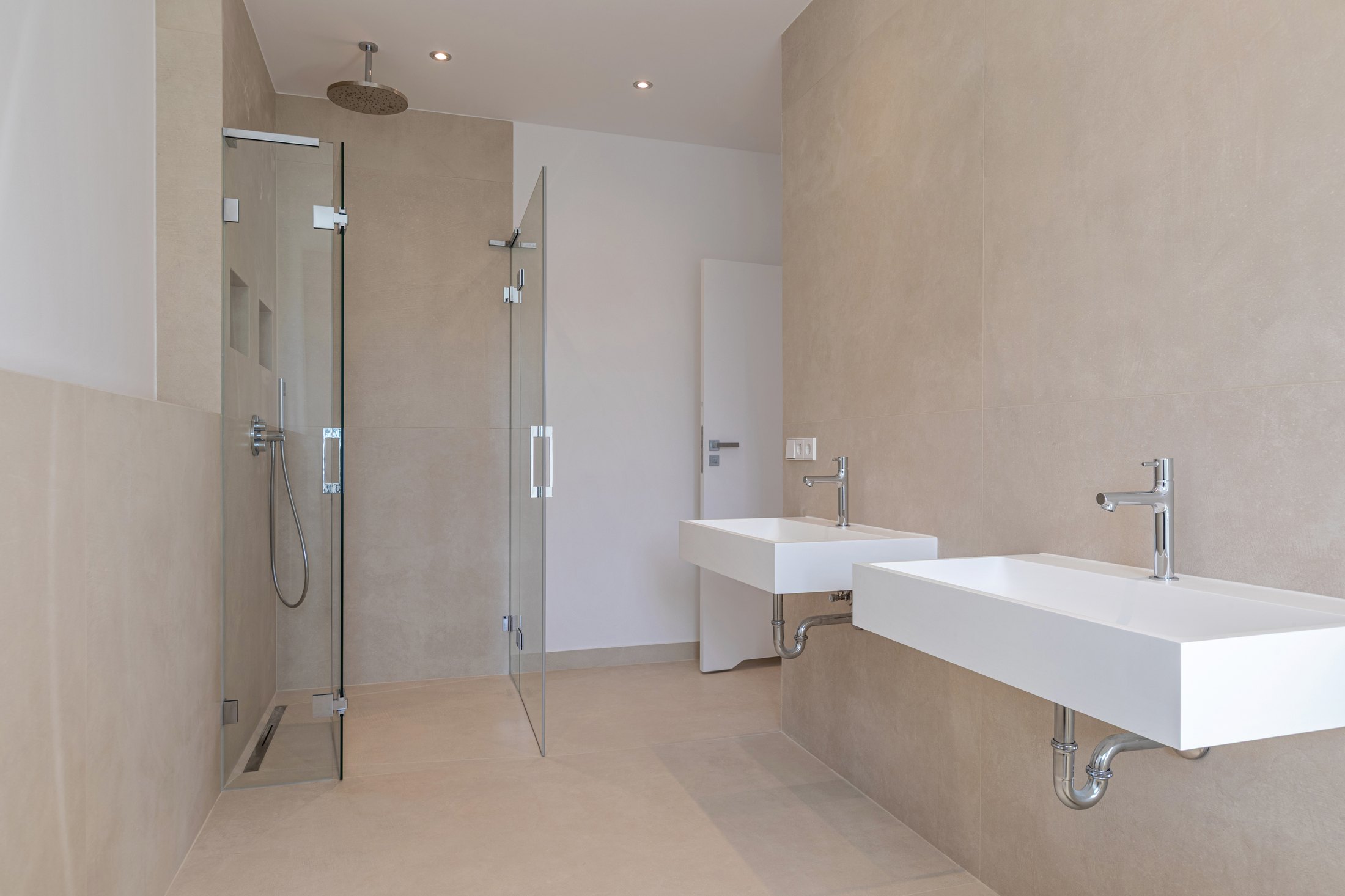 Minimalistisches, beiges Badezimmer mit begehbarer Glasdusche und zwei weißen Wandwaschbecken mit Chromarmaturen.