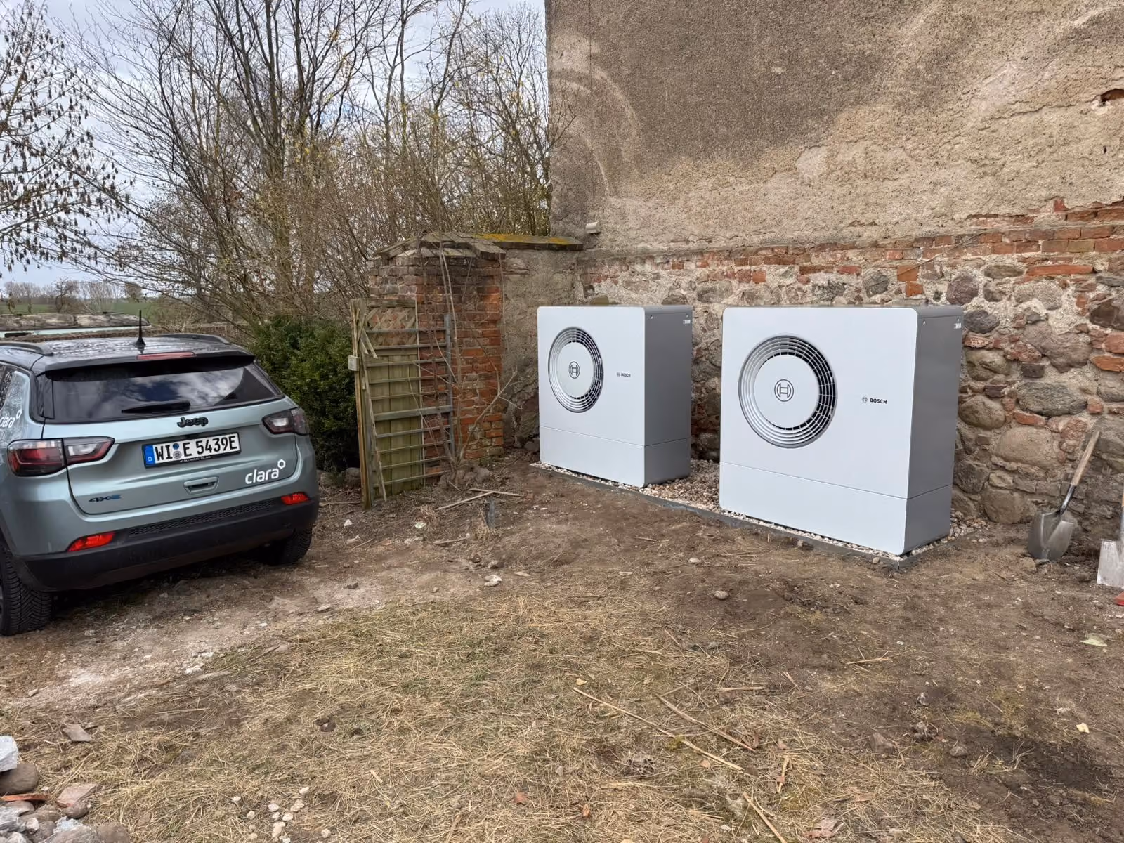 Zwei Bosch-Außenwärmepumpen, die auf Kies in der Nähe einer Steinmauer und neben einem geparkten Jeep aufgestellt sind.
