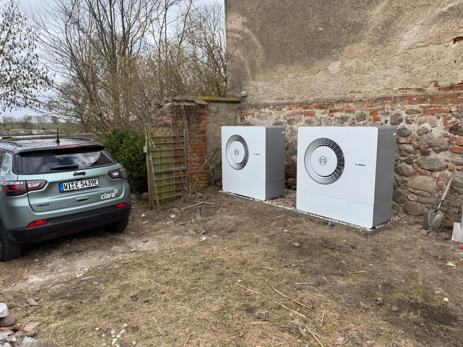 Zwei Bosch-Außenwärmepumpen, die auf Kies in der Nähe einer Steinmauer und neben einem geparkten Jeep aufgestellt sind.