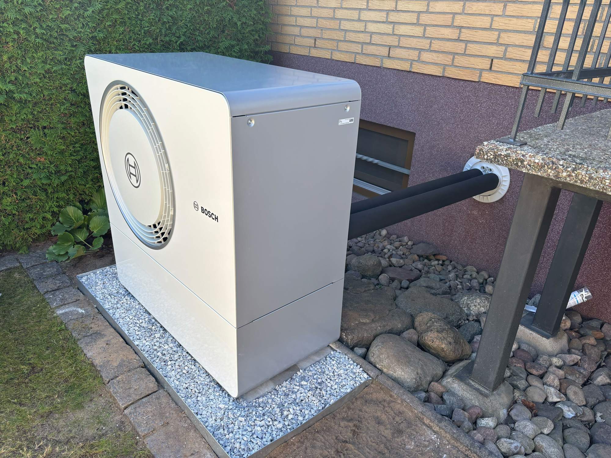 Ein Außenaggregat einer Bosch-Klimaanlage oder -Wärmepumpe, das auf Kies neben einer Hauswand aufgestellt ist.