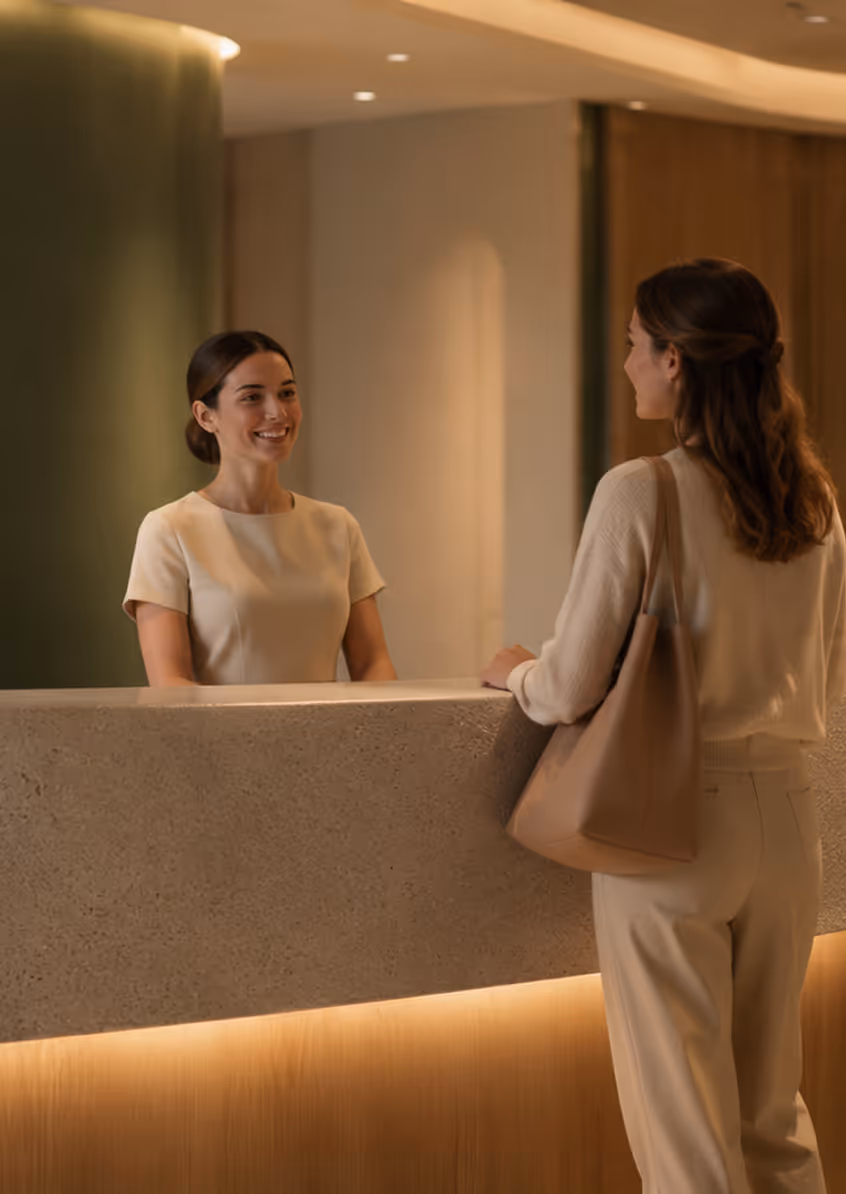 Smiling receptionist talking to a woman with a brown bag at a modern hotel front desk.