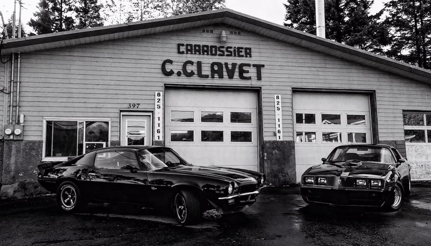 Black and white photo of two classic muscle cars parked outside Carrossier C. Clavet garage.