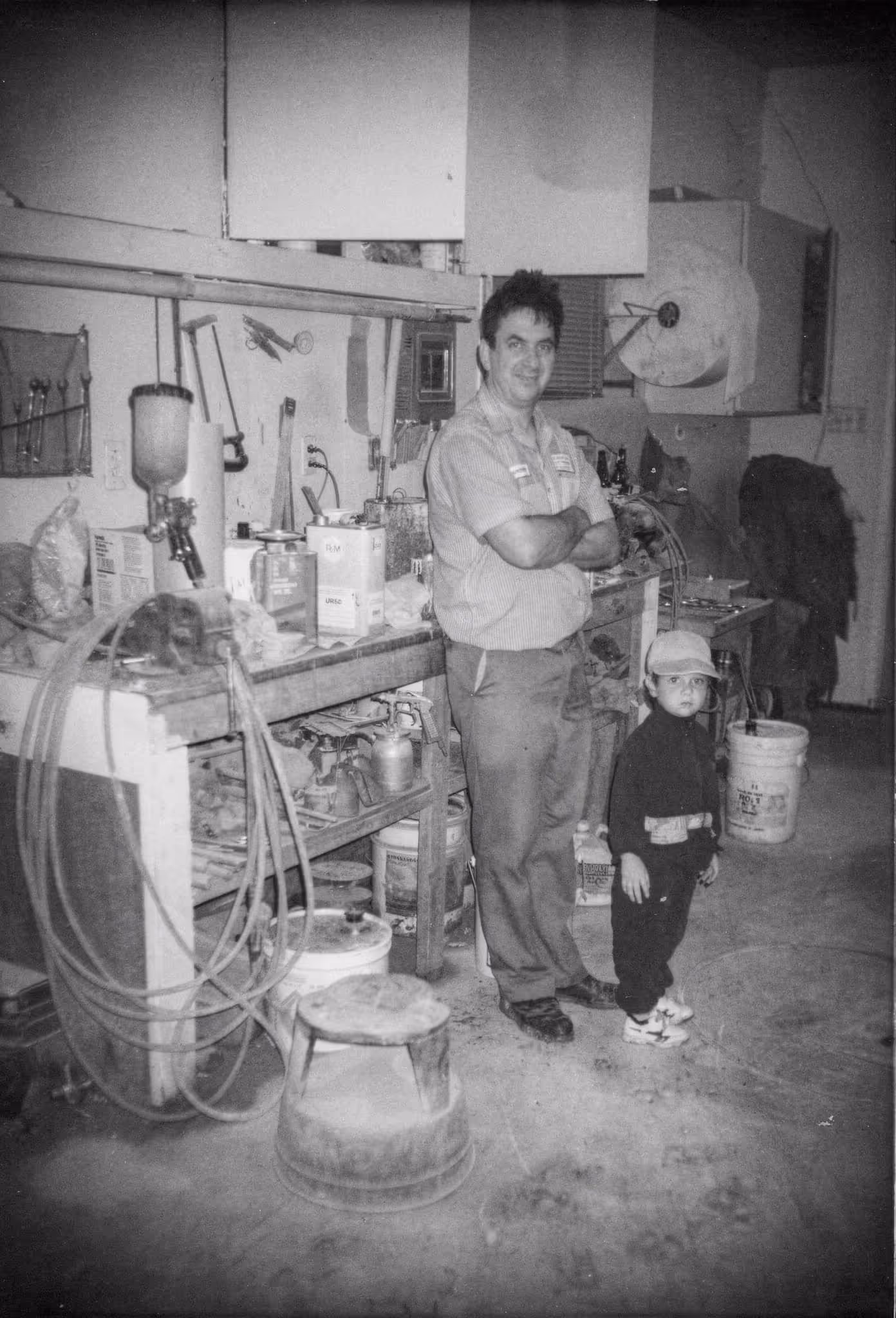 Black and white photo of a man standing with a child in a workshop surrounded by tools and equipment.