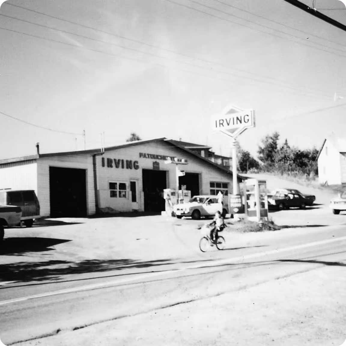 Black and white photo of an Irving gas station with cars and a person riding a bicycle nearby.