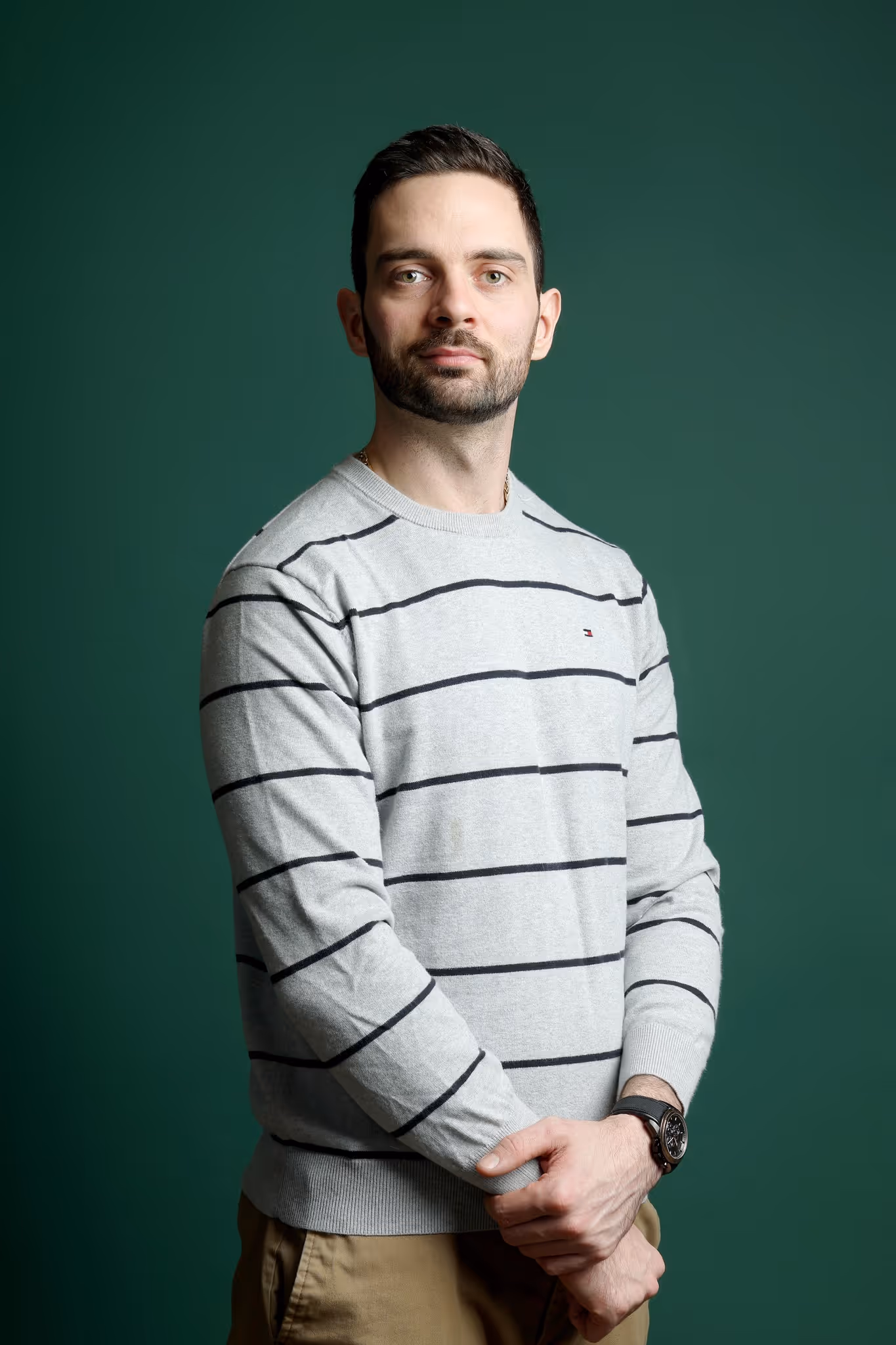 Man with beard wearing a gray striped sweater and watch, standing against a green background.