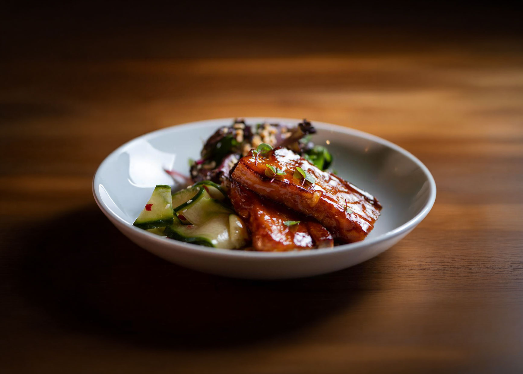 White bowl with glazed grilled tofu, cucumber salad, and mixed greens on a wooden table.