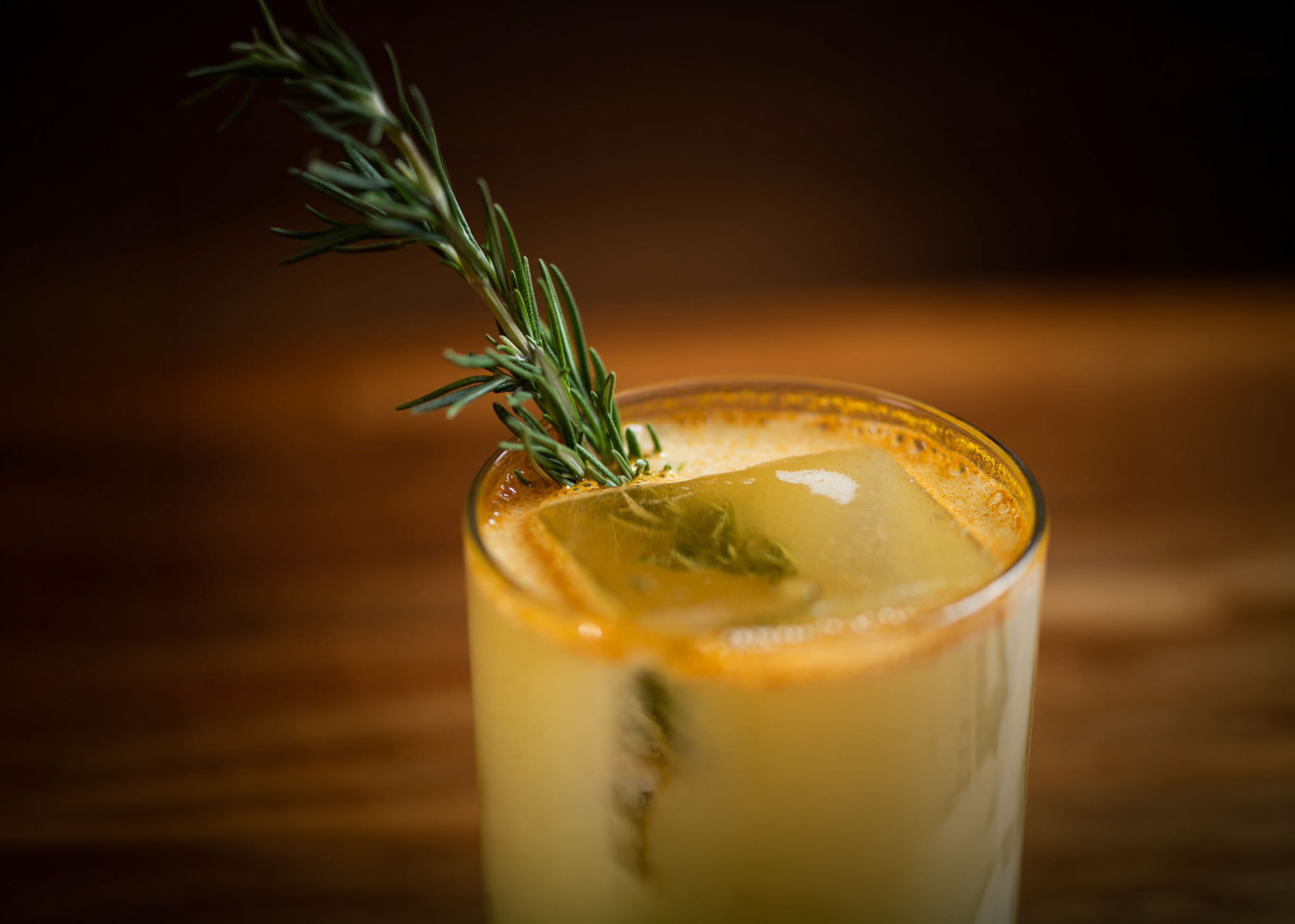 Close-up of a yellow cocktail garnished with a rosemary sprig and a large ice cube in a glass.