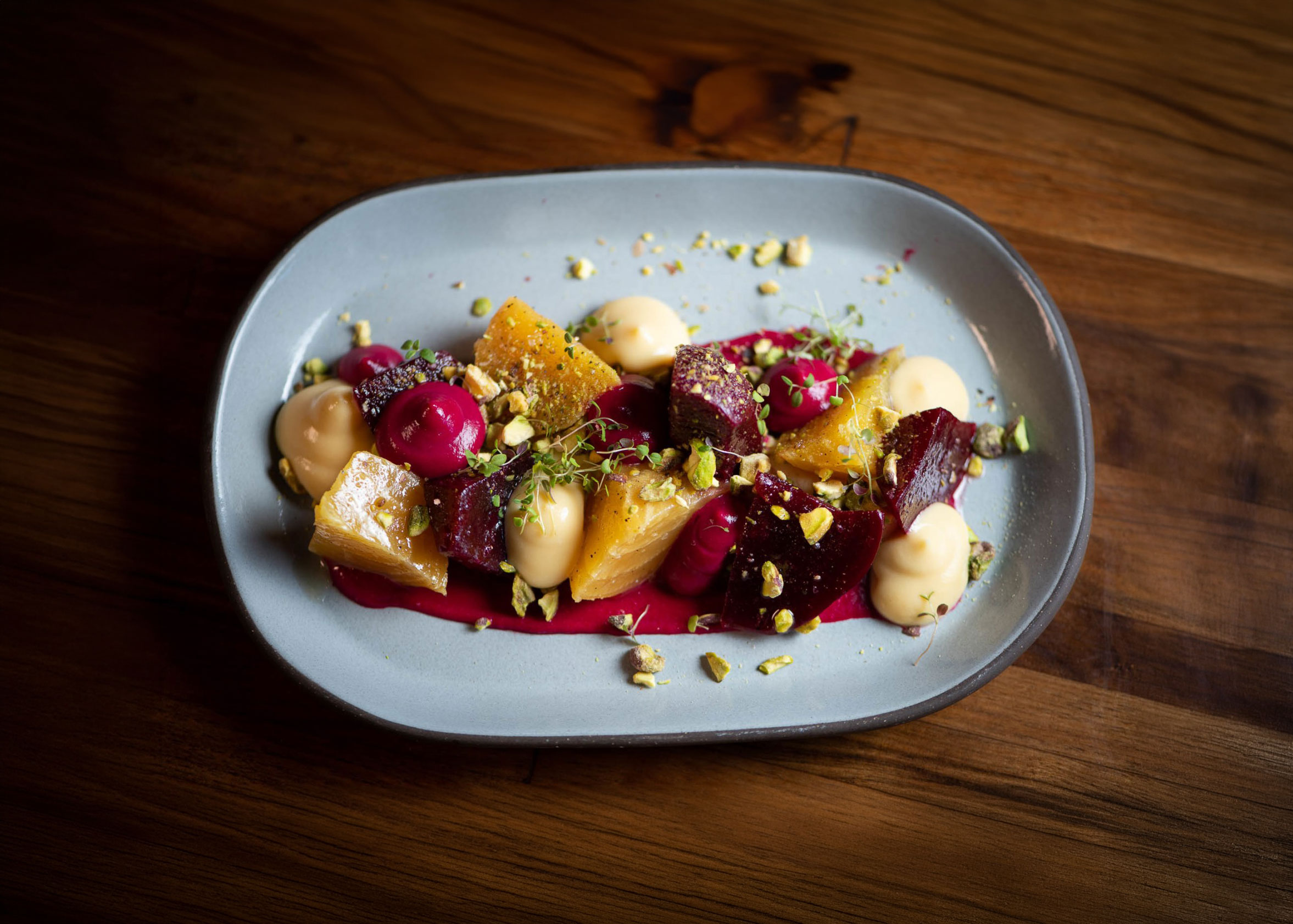 Plated gourmet dish with yellow and red beet pieces, dollops of sauce, pistachios, and microgreens on a gray plate.