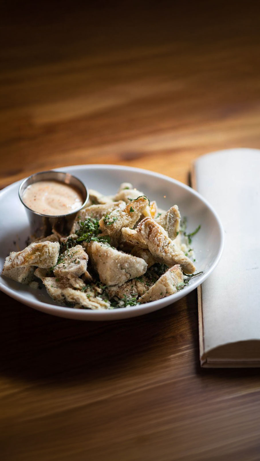 Fried artichoke hearts with herbs and dipping sauce in a white bowl on a wooden table.