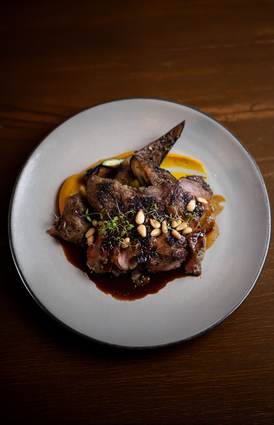 Plate with grilled meat slices topped with pine nuts and herbs, served with grilled vegetables and sauce.