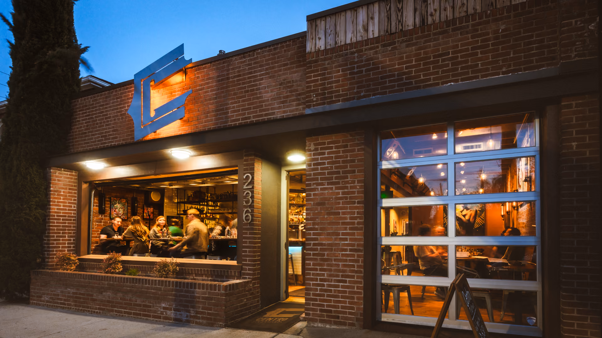 Evening view of a brick building with large open windows showing people dining inside a cozy restaurant with warm lighting.