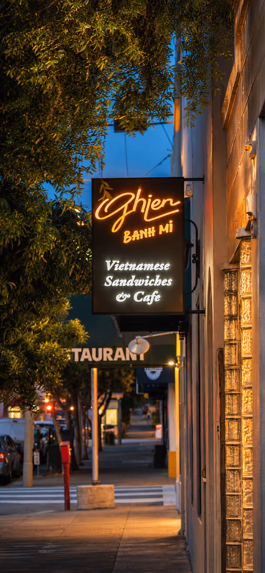 Glowing sign for Ghien Banh Mi Vietnamese Sandwiches & Cafe hanging on street during twilight.