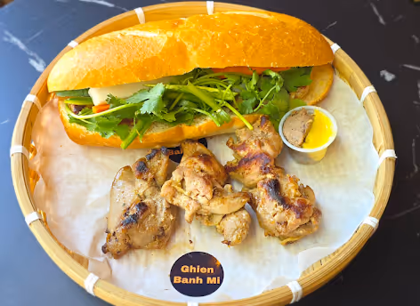 Banh mi sandwich with cilantro and grilled chicken pieces on bamboo tray with dipping sauce.