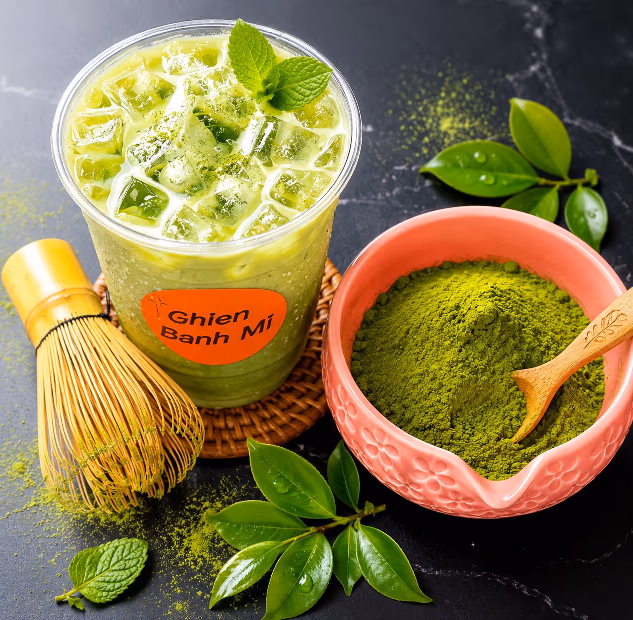 Iced matcha green tea in a plastic cup with a bamboo whisk and matcha powder in a pink bowl with spoon.