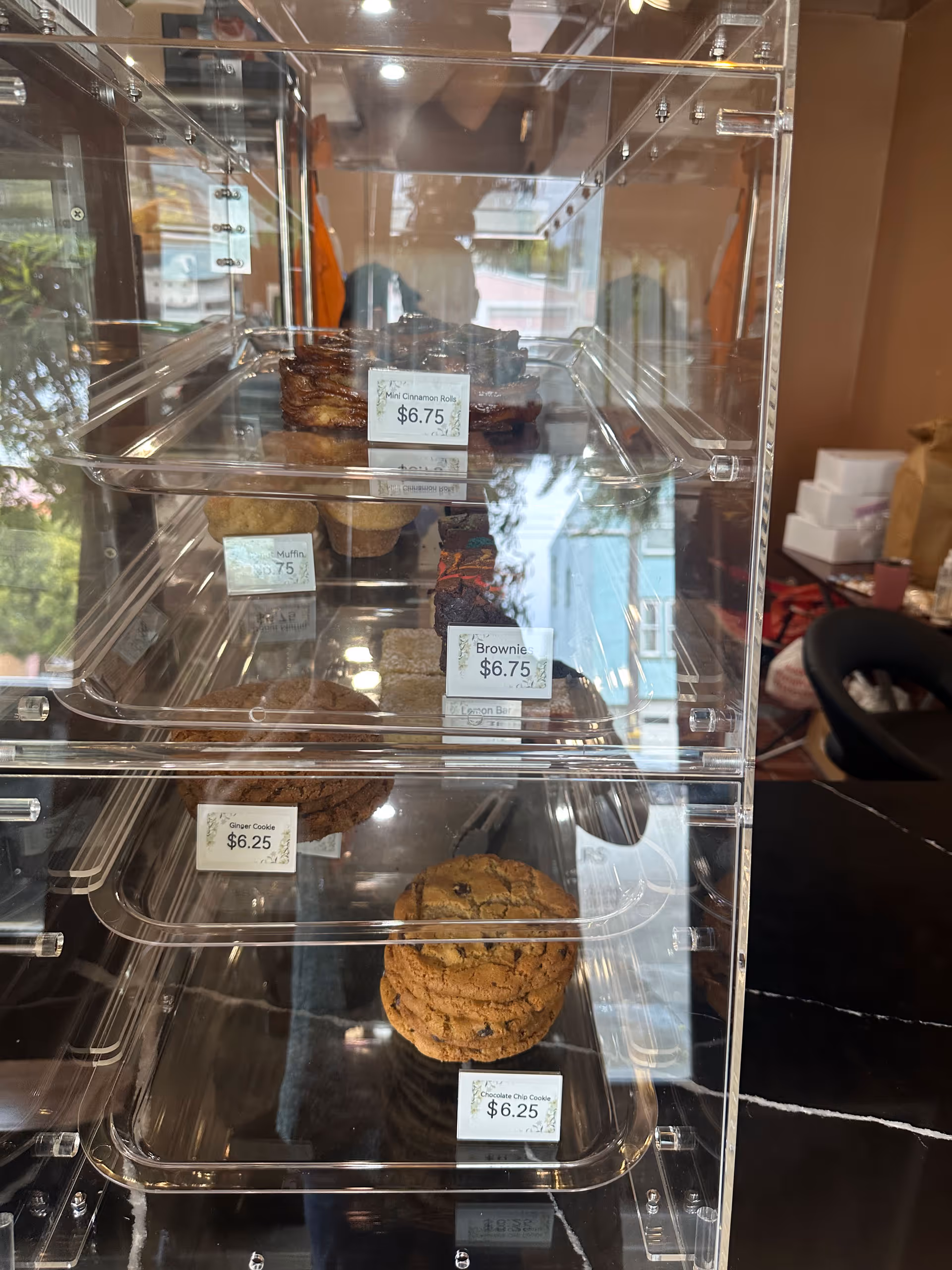 Clear display case with mini cinnamon rolls, muffins, brownies, ginger cookies, and chocolate chip cookies with price tags.