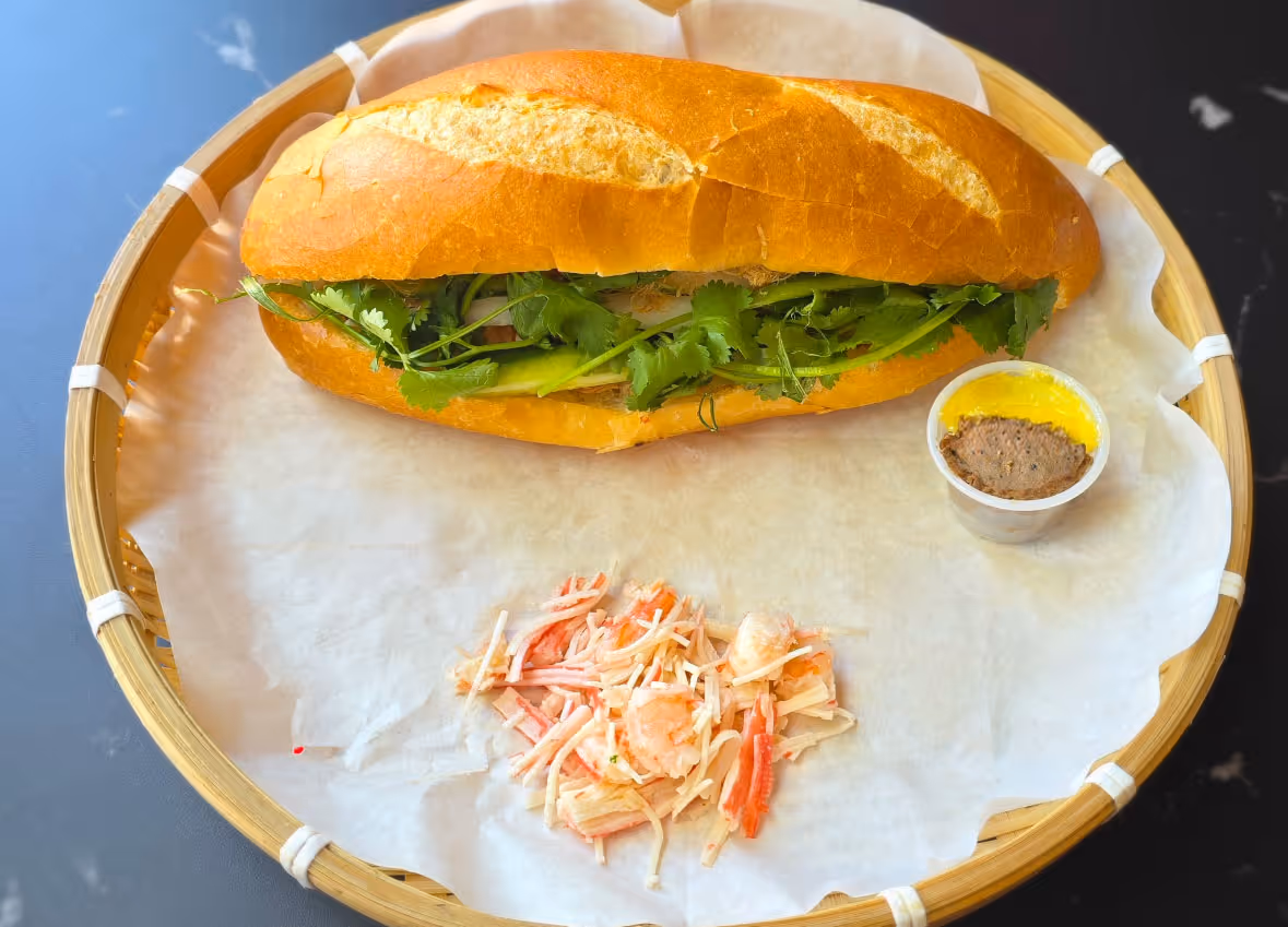 Sandwich with fresh cilantro on a bamboo tray with a small cup of pâté and shredded salad.