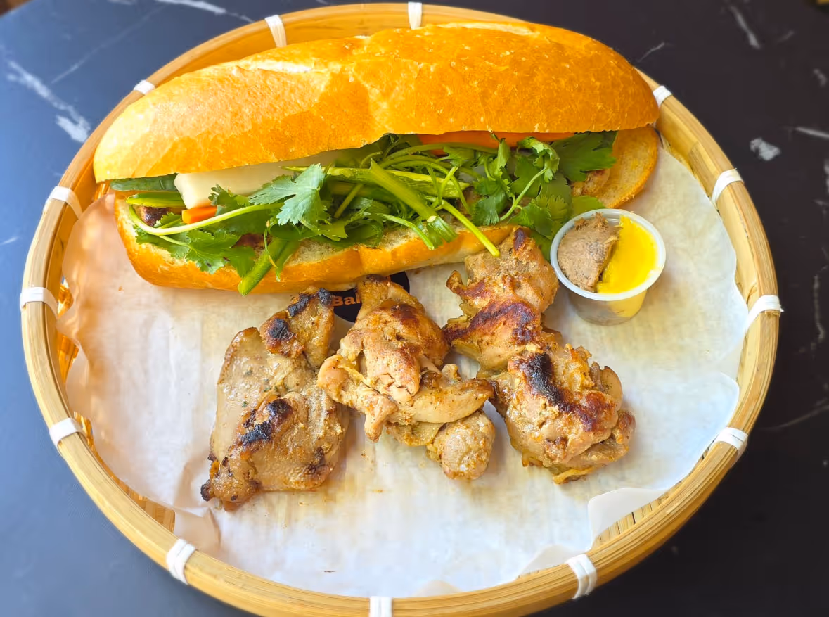 Grilled chicken pieces with sandwich bun filled with herbs and vegetables served in a bamboo basket.