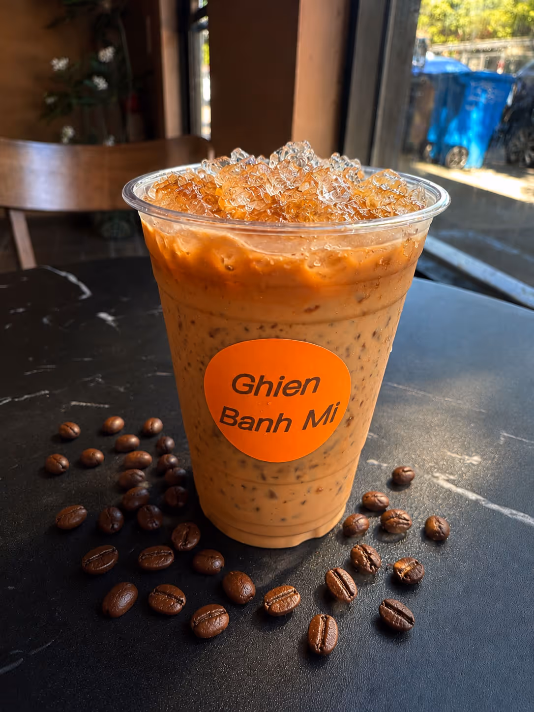 Iced coffee in plastic cup labeled Ghien Banh Mi with coffee beans scattered on a black table.