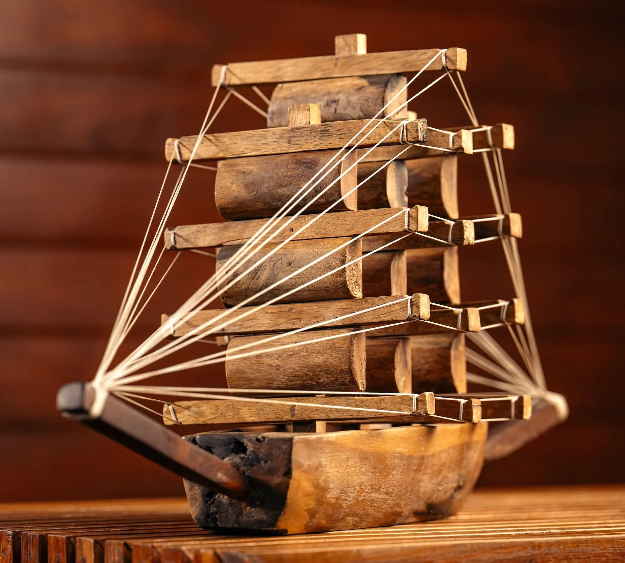 Wooden model of a sailboat with string rigging on a wooden surface and brown background.